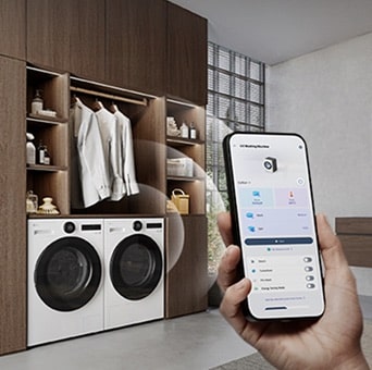 An LG washer and dryer set is built into the bottom of a wooden wardrobe with clothes and accessories. A hand holds a smartphone running the LG ThinQ app in front.
