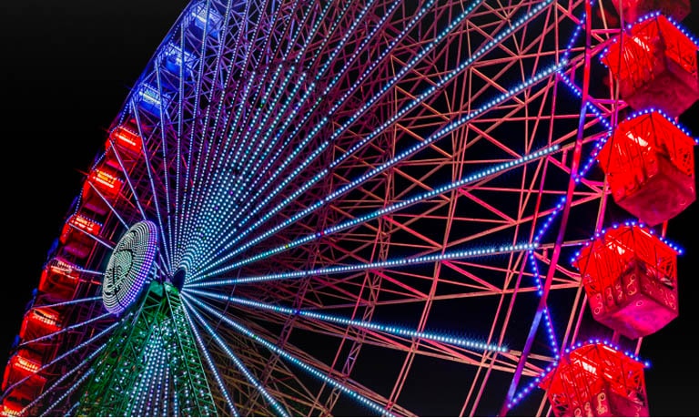 Ein hell erleuchtetes, regenbogenfarbenes Riesenrad vor einem schwarzen Nachthimmel, aufgenommen aus einem niedrigen Winkel.