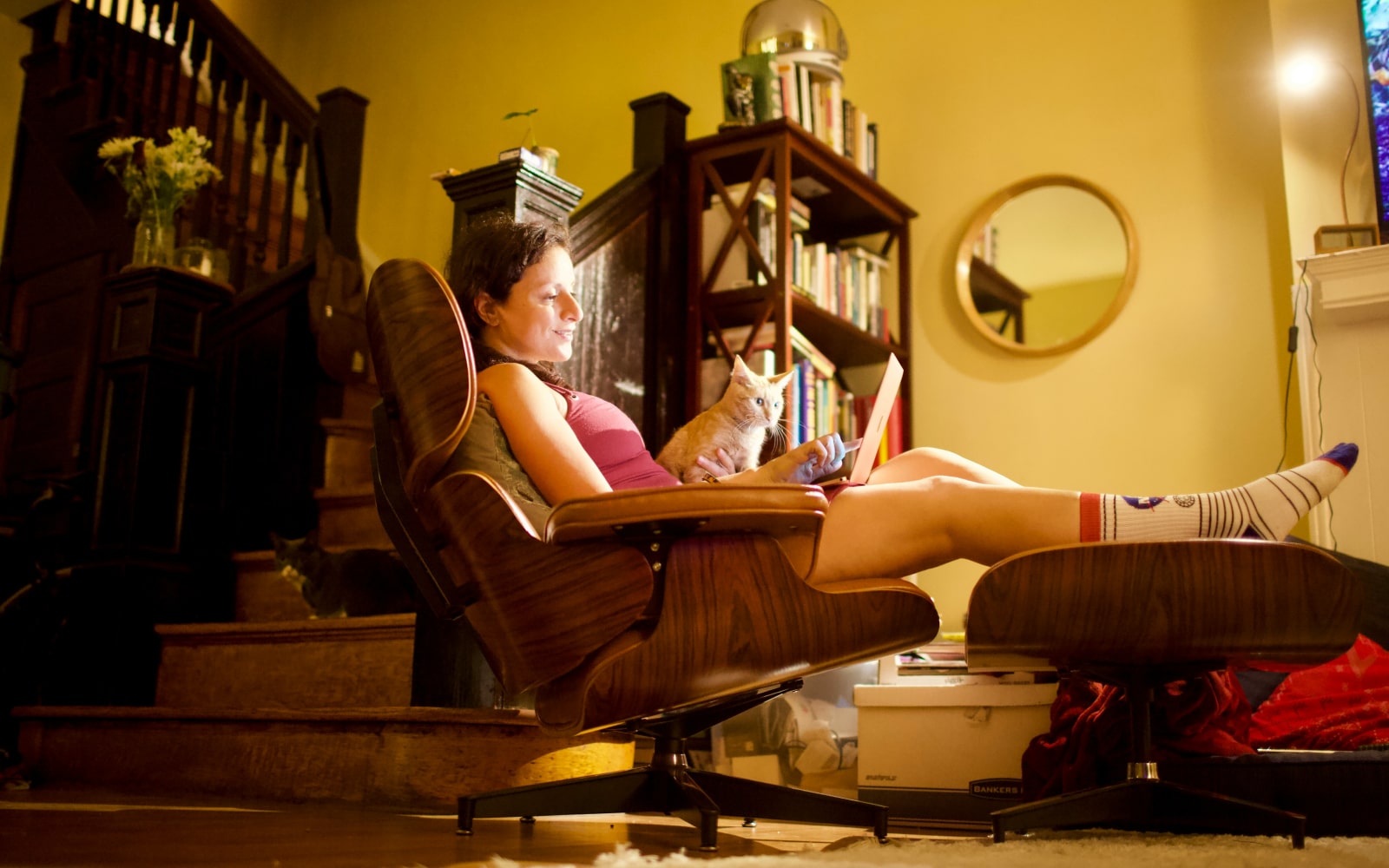 Woman writing on her laptop in a recliner chair in a cosy living room, with soft lighting and a television glowing in the background.