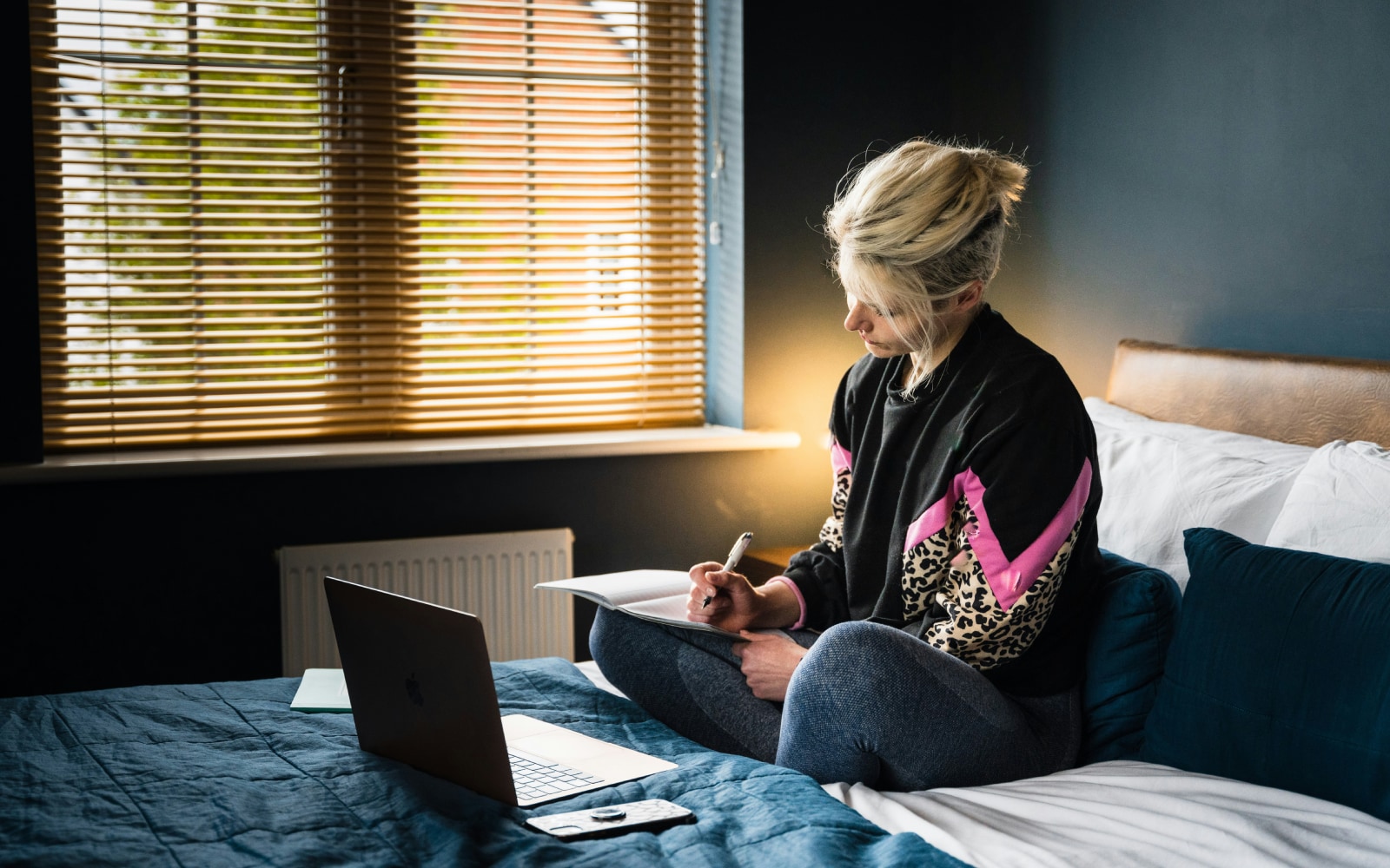 A woman writing in a notebook with a laptop open in front of her on a bed, in a room with natural light coming through venetian blinds.