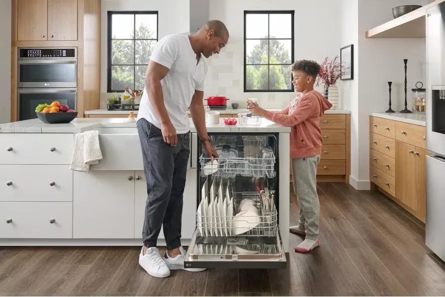  A man and a young boy are unloading dishes from a dishwasher in a bright kitchen with white and wooden cabinets. The man is taking out a plate while the boy is holding a glass. The kitchen is spacious with a large island in the centre.