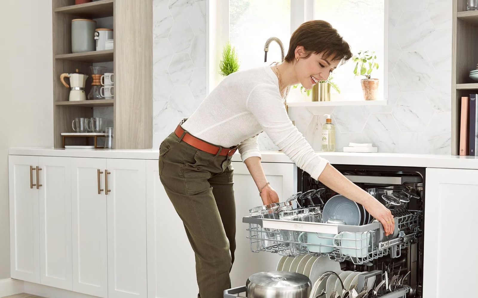 A woman is loading a dishwasher in a modern kitchen with white cabinets and a patterned rug on the floor. The dishwasher is fully open, and she is placing a dish on the top rack. 