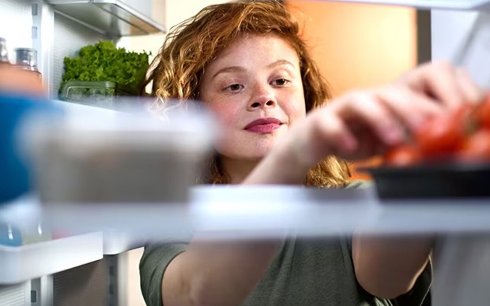 The view is from inside the fridge, looking out. Someone is reaching inside the fridge for tomatoes.