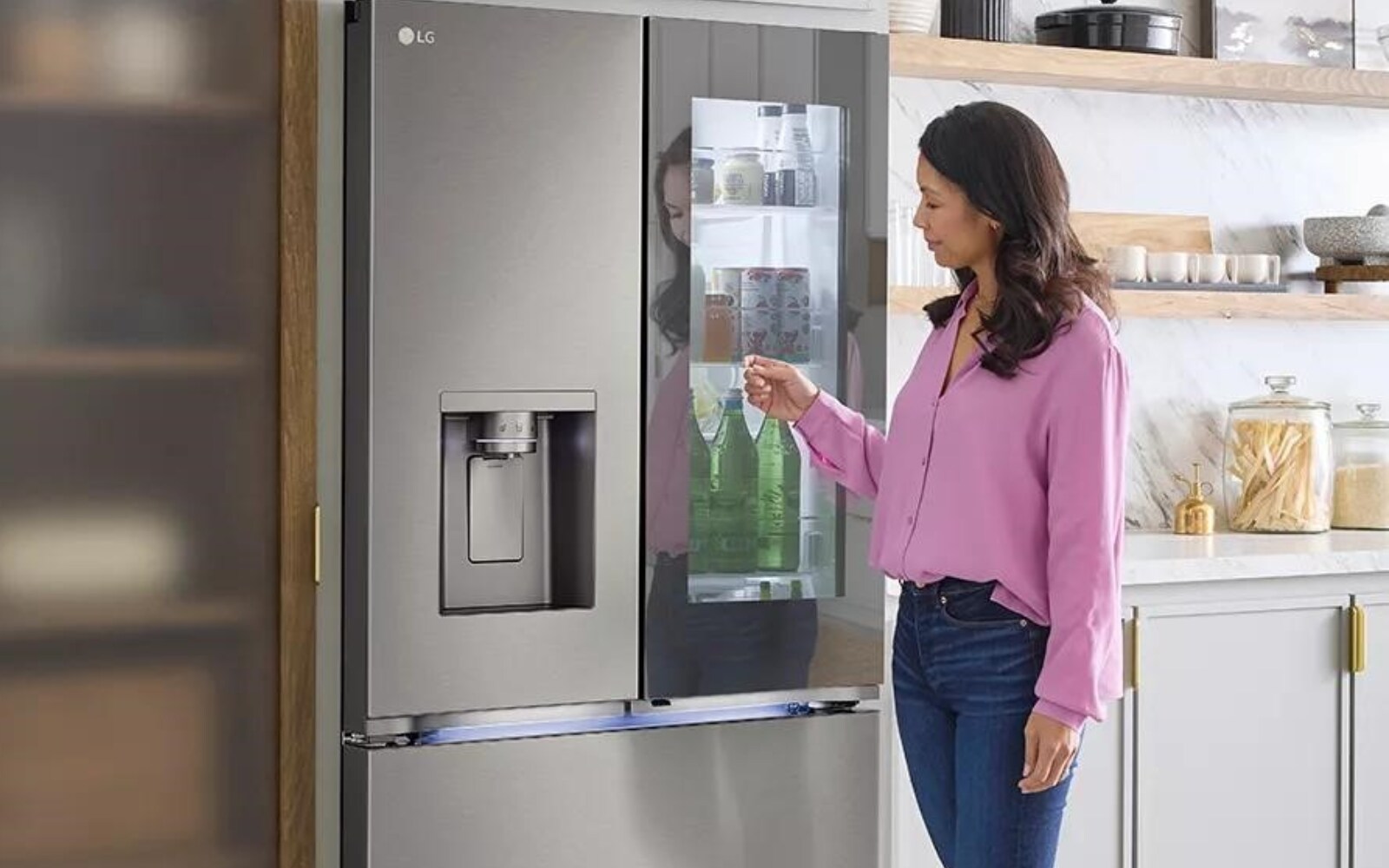 : A woman knocking on the InstaView glass door on the modern stainless steel LG French door refrigerator. She is accessing the easy-access compartment where drinks and frequently used items are stored, while the refrigerator also features a built-in water and ice dispenser on the left door.