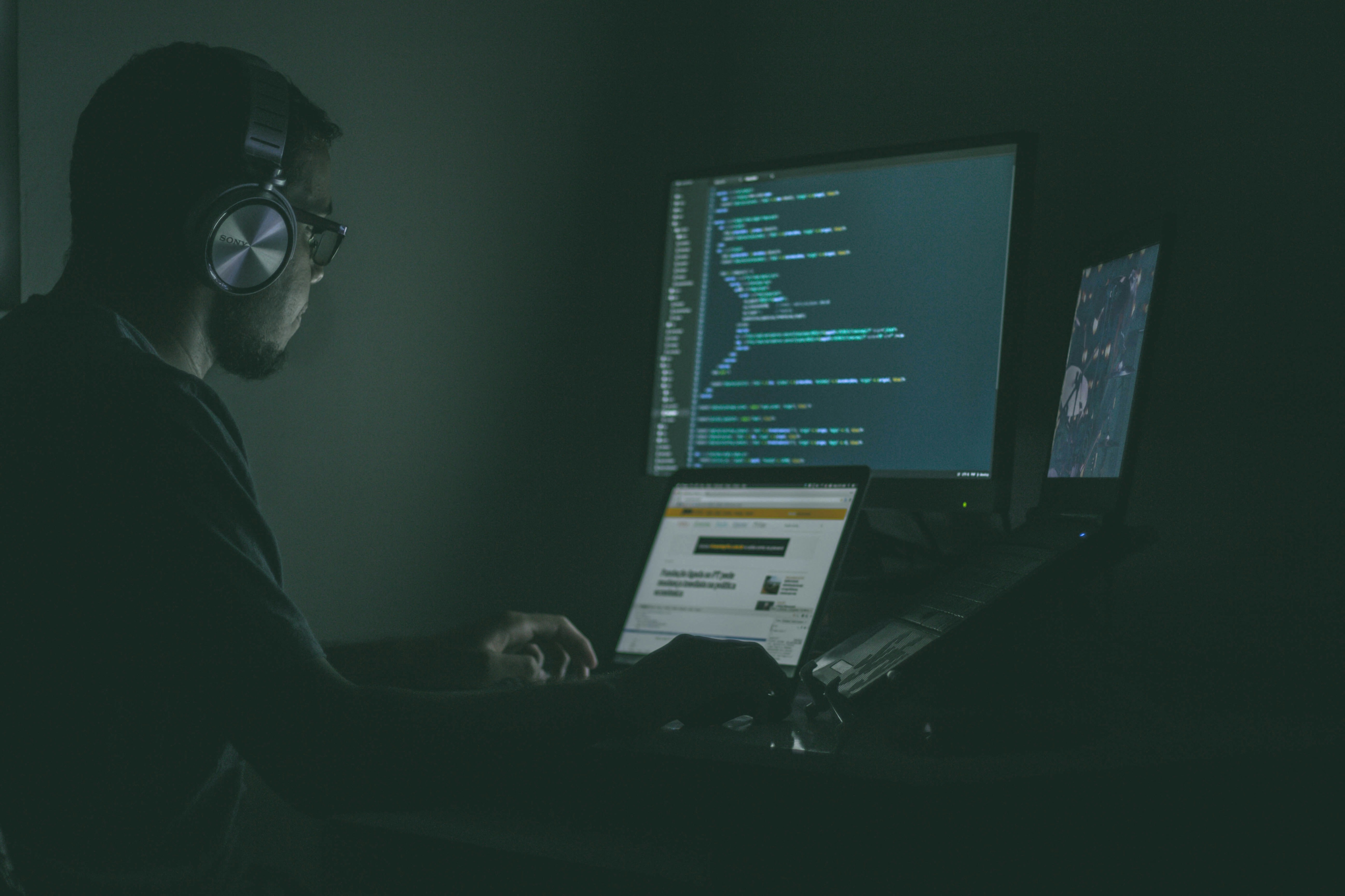 Man wearing headphones working on a laptop and an additional monitor displaying lines of code in a dimly lit room.