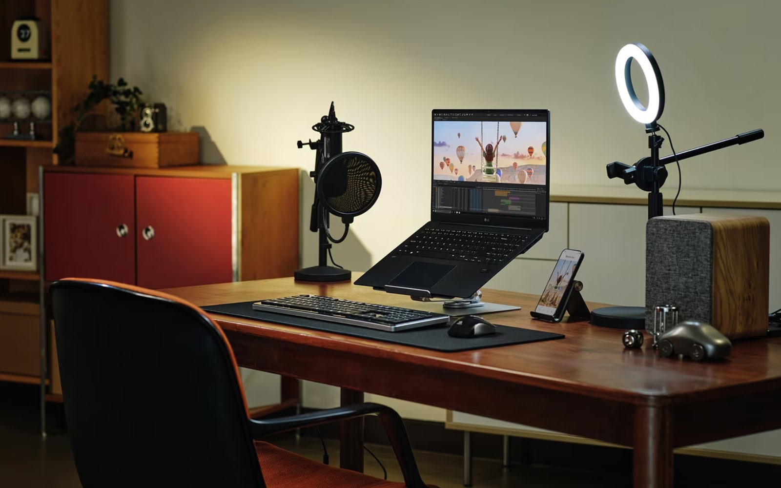 A home office setup with a LG gram Laptop, keyboard, mouse, microphone, and ring light on a wooden desk. Shelves and a cabinet are in the background.