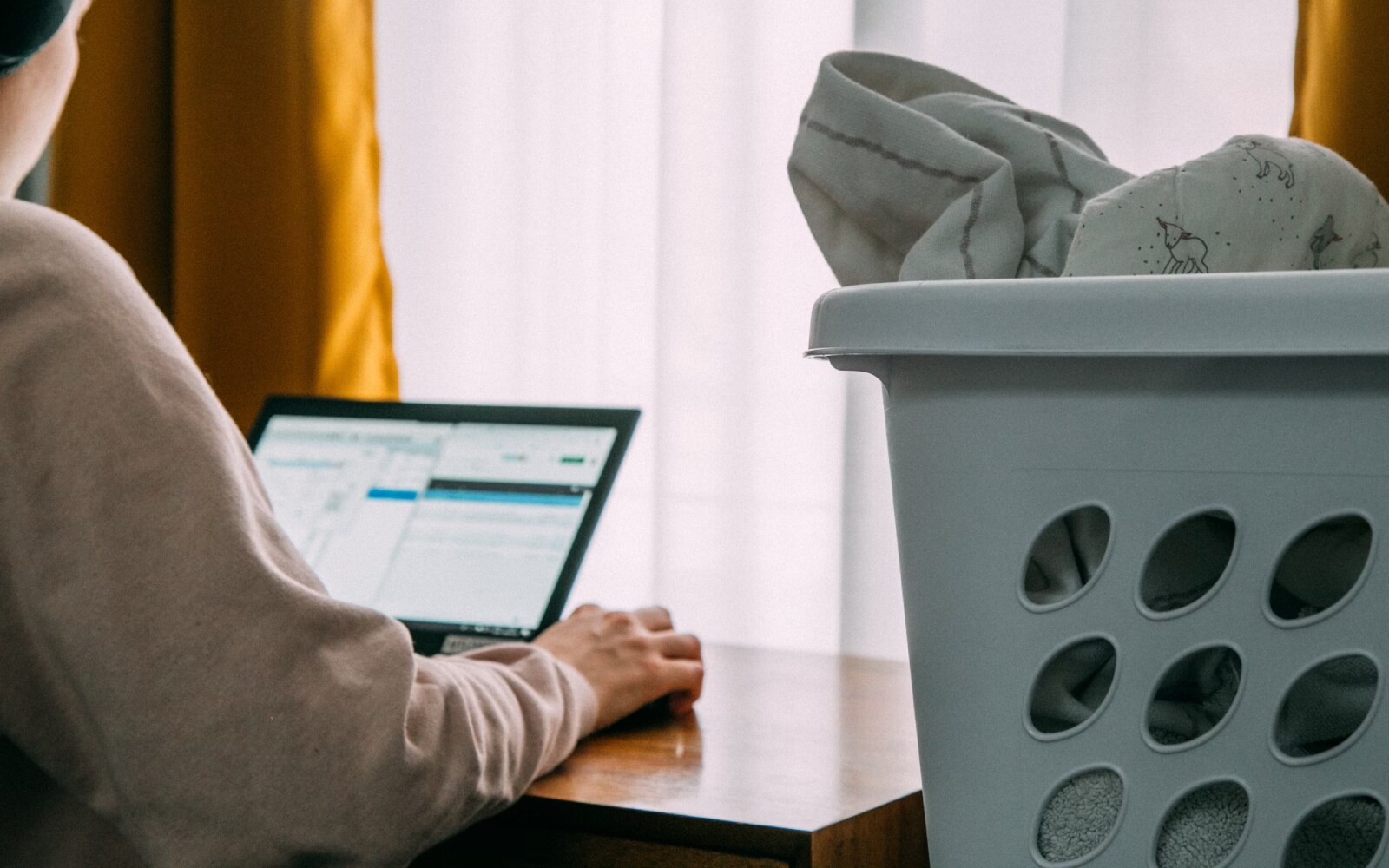 Person using a laptop next to a window with yellow curtains, while a laundry basket filled with clothes is in the foreground. 