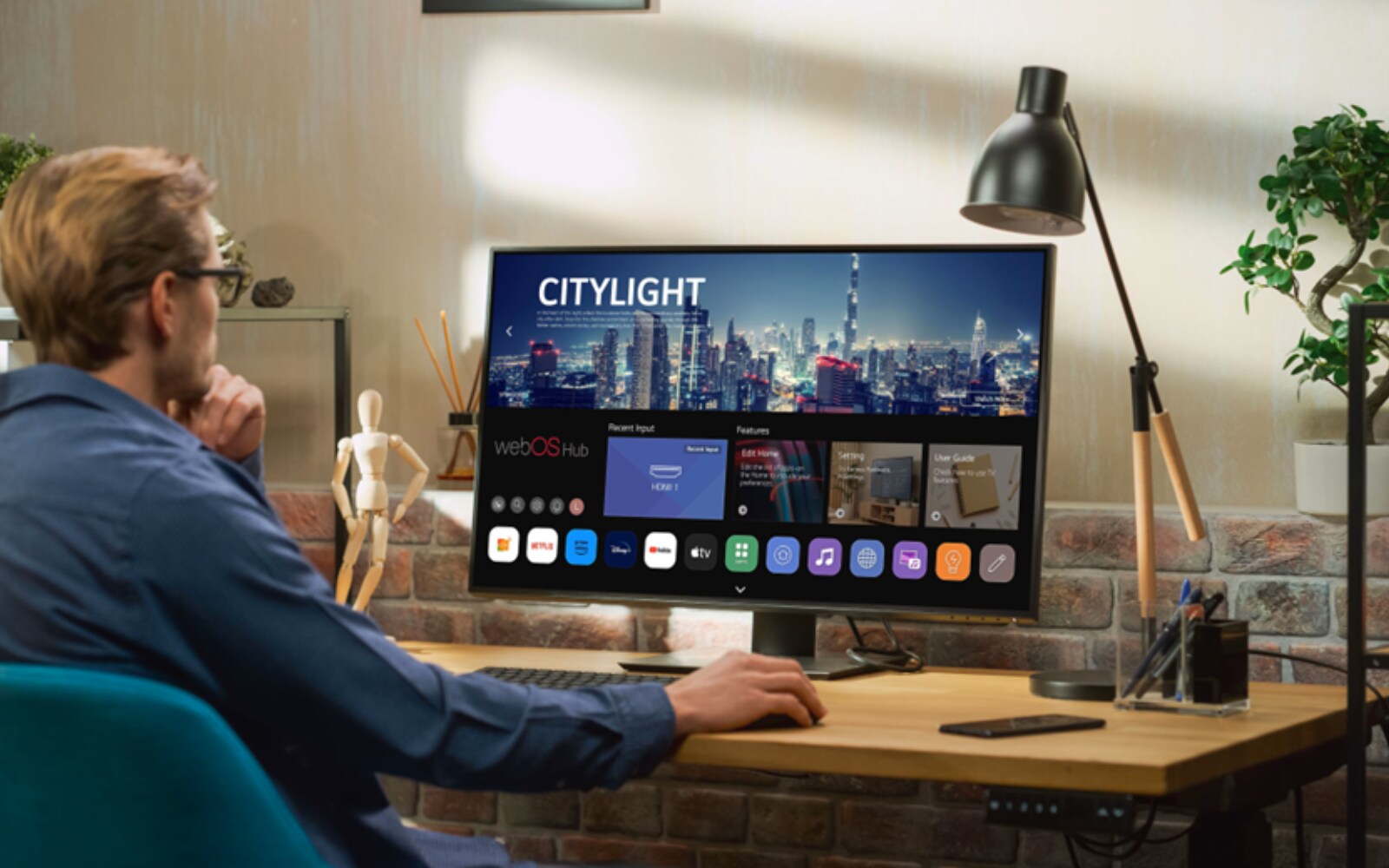 person seated at a desk using an LG Smart Monitor displaying a LG webOS interface with various apps. The workspace includes a lamp, plants, and a modern decor with a brick wall, creating a cozy office setting.