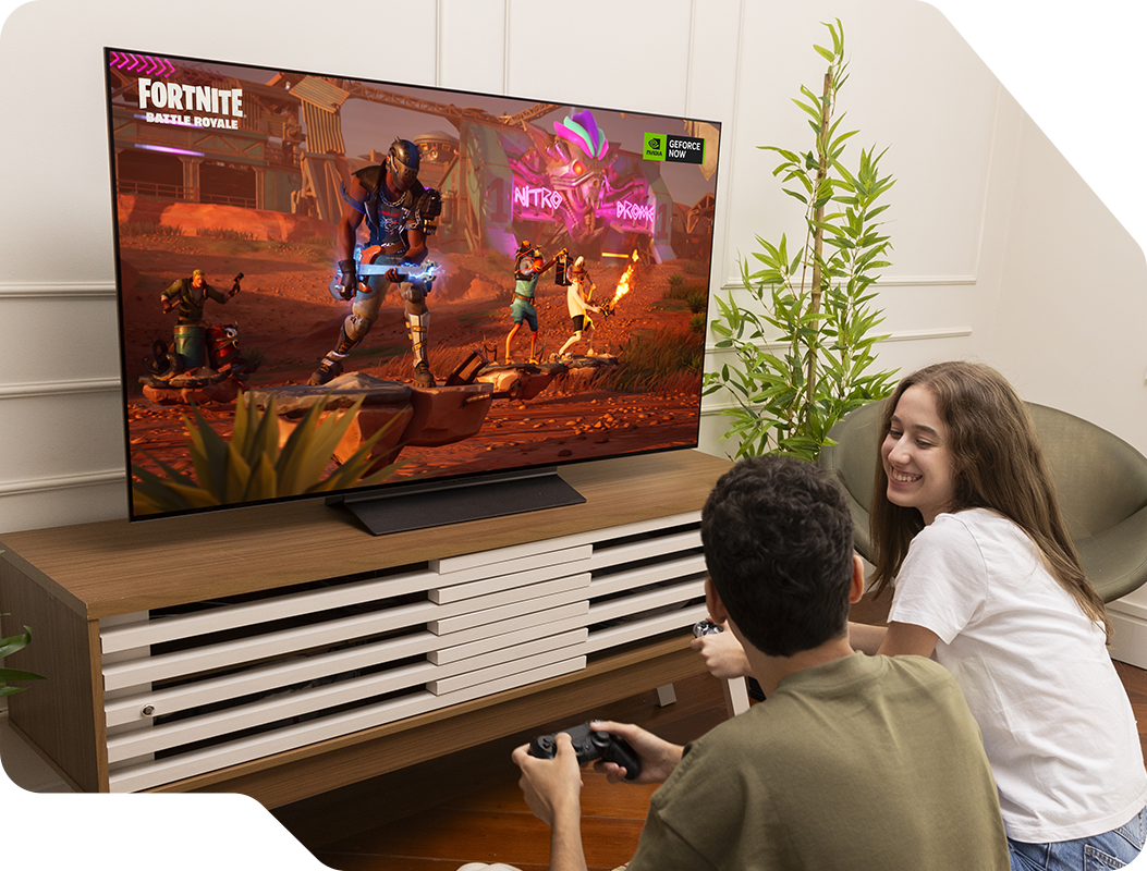 Bruno e sua namorada jogando FORTNITE na TV LG OLED C4 na sala de estar. Sua namorada sorri para Bruno. Planta ao lado direito da TV.