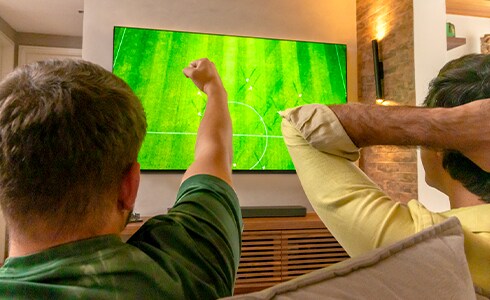 Dois amigos torcendo, um com o punho alto e o outro segurando a cabeça. Eles estão olhando para a tela do campo de futebol na TV LG OLED.