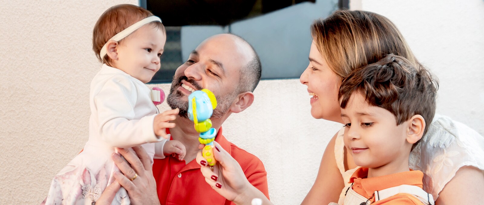 Um corte familiar, a mãe a dar um brinquedo à filha, o pai a segurar a filha a sorrir. Filho olhando para baixo com prazer na cara.