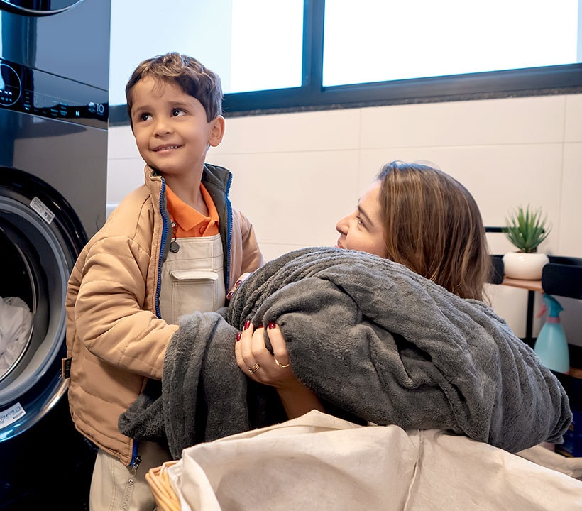 Mãe e filho a segurar outra roupa suja, um cobertor escuro grande. Filho sorridente, alegria de ajudar a mãe. A mãe olha para o filho com alegria.