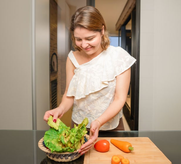 A mãe prepara-se para a refeição, manipulando ingredientes. Alguns vegetais verdes frescos, tomate e cenouras. Ela está a usar camisas brancas.