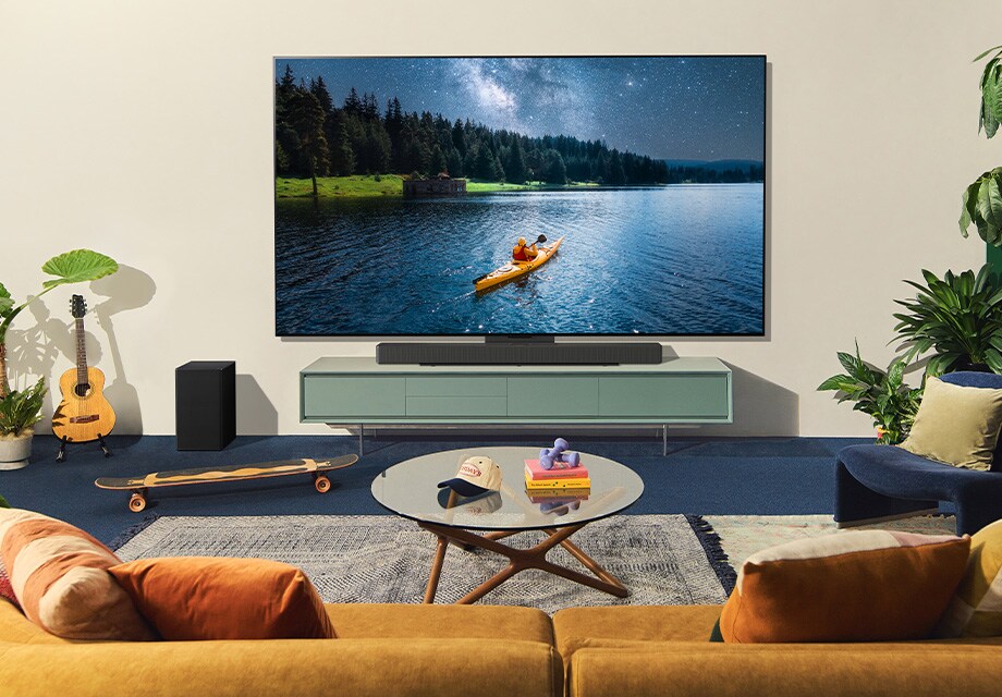 Sala de estar aconchegante com duas pessoas assistindo a um caiaqueiro na LG OLED TV na parede, com plantas, guitarra, livros e móveis aconchegantes