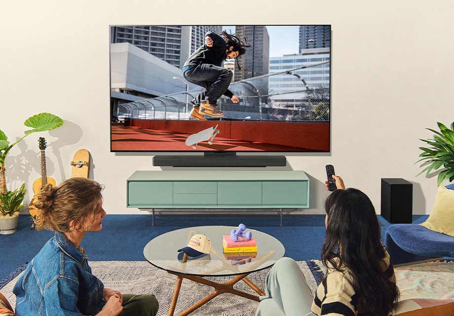 Sala de estar com duas pessoas assistindo a um skatista na LG OLED TV montada na parede, rodeada de plantas, livros e decoração moderna