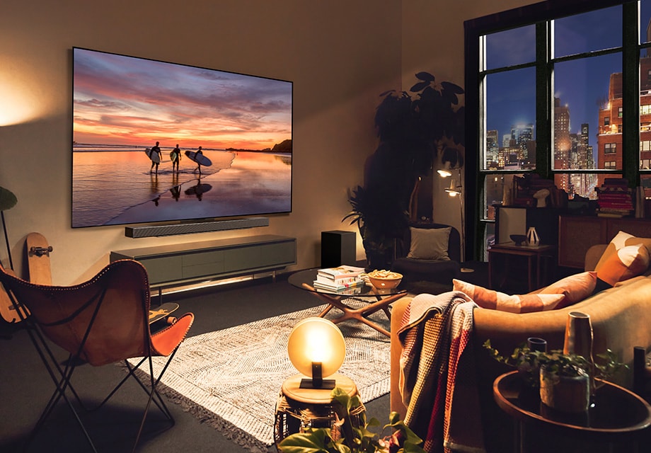 Sala de estar com iluminação ambiente e LG OLED TV mostrando uma cena de pôr do sol na praia, com luzes quentes, plantas e decoração moderna