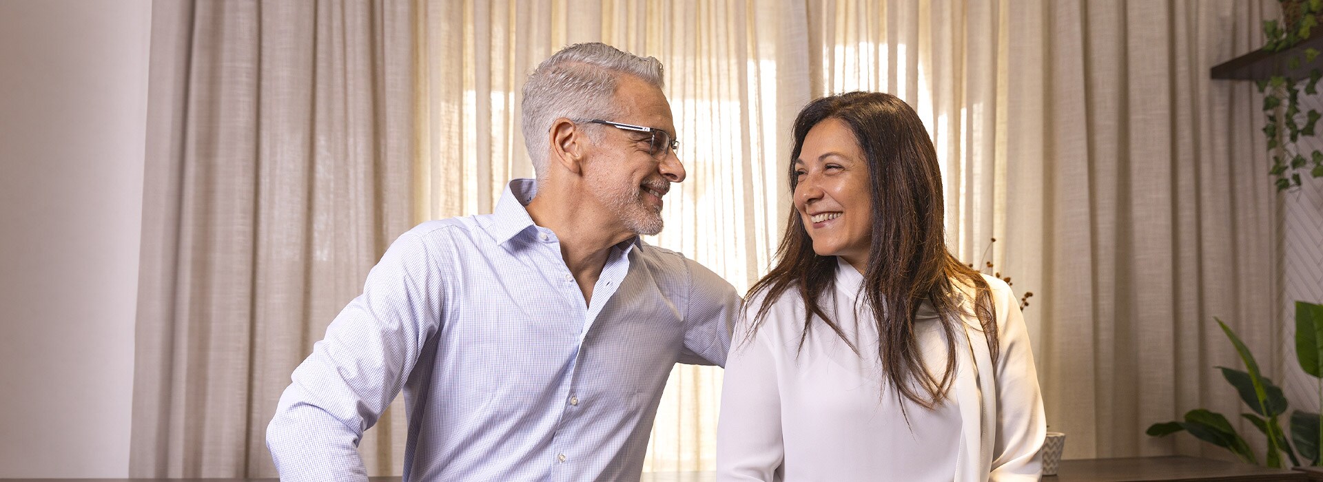 Luis e Rosana sorrindo um para o outro, sentados em frente a uma cortina.