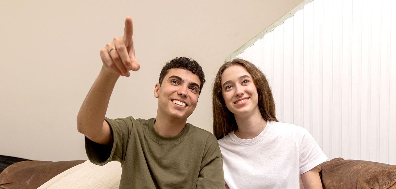 Dois jovens sorrindo e fazendo um gesto enquanto estão sentados no sofá.