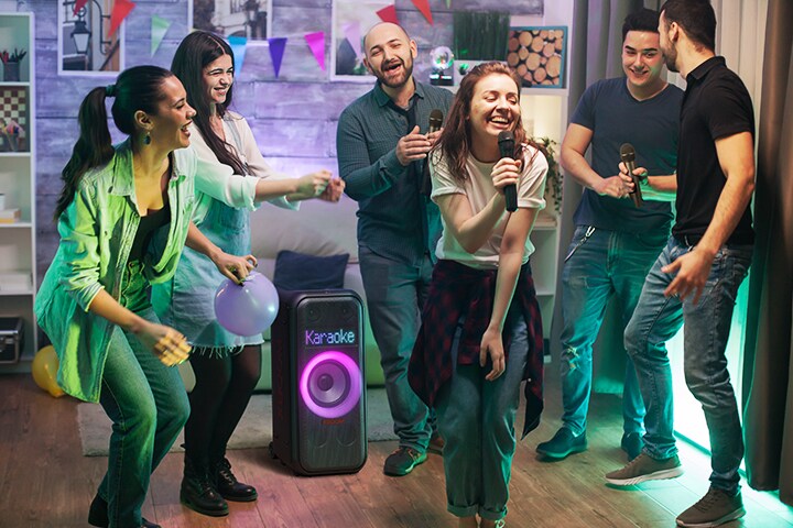 Há pessoas curtindo karaokê na sala de estar. Entre elas, o XL7T está colocado no chão com iluminação woofer ligada. No painel de LED, a palavra "Karaoke" é exibida.