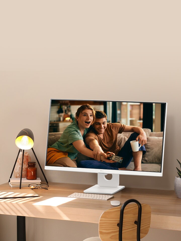 Na mesa perto da janela iluminada pelo sol, há um monitor inteligente exibindo um filme em sua tela. Ao lado do monitor, há uma lâmpada e um vaso de plantas. Na mesa, há um teclado e um mouse.