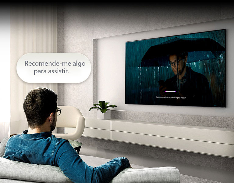 Um homem sentado no sofá olha para a TV na parede oposta. Um balão de fala com os dizeres “Recomende-me algo para assistir” flutua acima da cabeça dele. Na tela, o vídeo de um homem usando guarda-chuva é reproduzido com uma IU que aceita reconhecimento de voz.