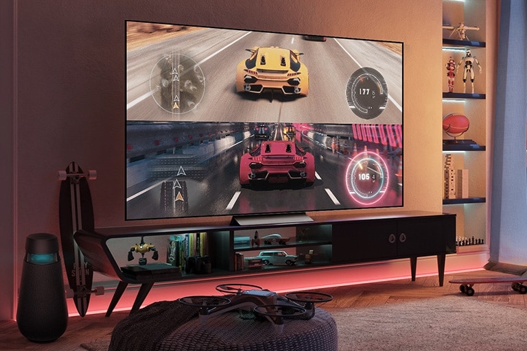 Em uma sala de jogos com iluminação avermelhada, um game de corrida automobilística com carros amarelo e vermelho competindo é exibida numa grande tela de TV. No lado esquerdo, há um alto-falante gamer dedicado, além de acessórios relacionados a jogos e outros dispositivos espalhados.