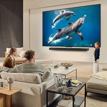 A family sits on a couch facing a LG QNED TV mounted on a wall above a LG Soundbar with a little girl pointing to a screen depicting two dolphins. The title talks about how every action is more thrilling on a massive screen.