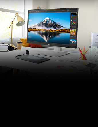On the desk, there is an UltraFine monitor displaying Photoshop work. Next to the monitor are a desk lamp, a mouse, paper, and coloured pencils.
