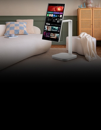 An LG Smart Monitor is placed in a living room in portrait mode, displaying various content apps. The monitor is mounted on a rotatable stand, surrounded by cosy interior elements such as a sofa, cushions, and a cabinet in the background.