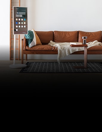 LG StanbyME, a vertical smart display, stands next to a leather sofa in a modern living room, showing app icons and widgets on the screen.