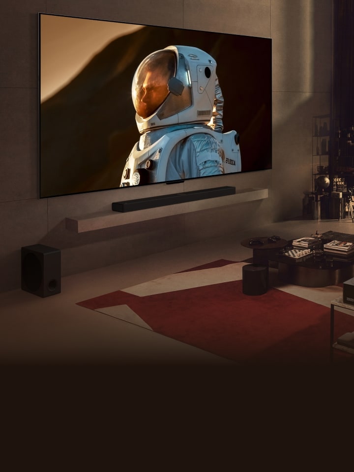 An ultra-large LG TV mounted on a wall in a cozy living room displaying a close-up shot of astronaut takes up the majority of the wall. A man sits on a sofa in front watching the TV with a dog, and a night cityscape is beyond the window.