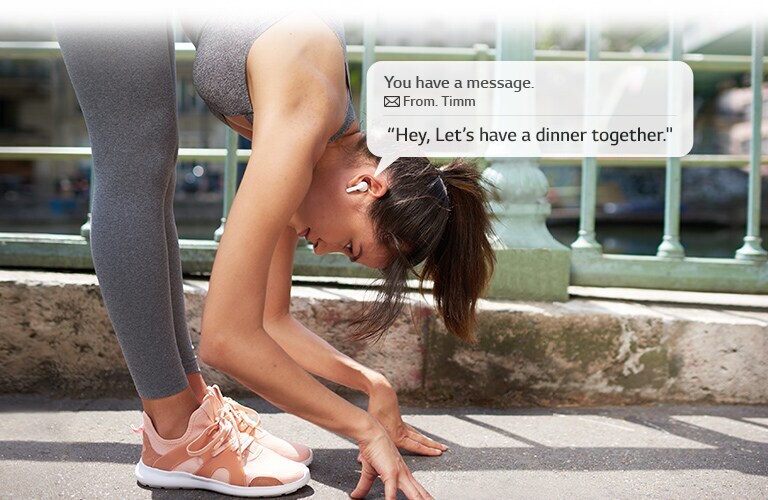 A woman is stretching and leaning down to touch the ground while she listens to a message that her earbuds read for her.