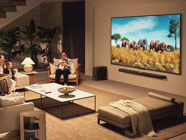 Une famille est assise sur un canapé face à un LG QNED TV monté sur un mur au-dessus d’une LG Soundbar dans un salon moderne.  La boîte Zero Connect se trouve à l’intérieur d’une table basse.