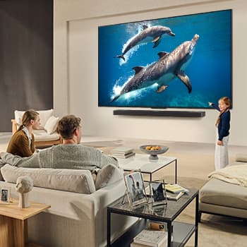 Une famille est assise sur un canapé face à un LG QNED TV monté sur un mur au-dessus d’un LG Soundbar. Une petite fille pointe du doigt un écran représentant deux dauphins. Le titre met en avant comment chaque action devient encore plus captivante sur un écran géant.