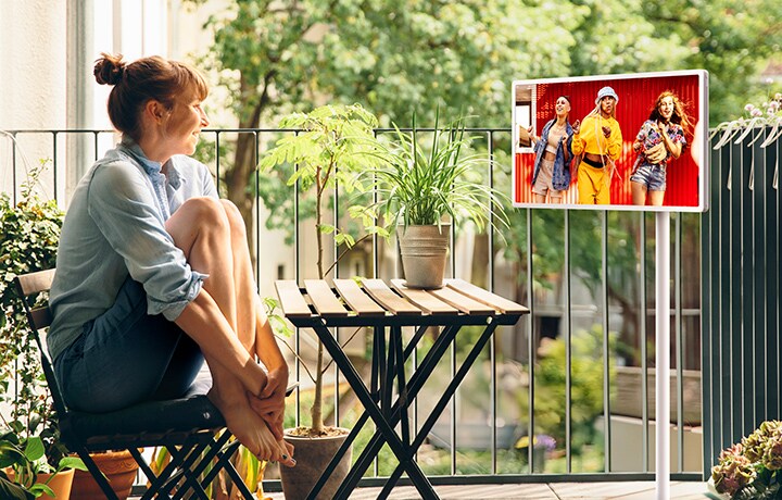 Une femme assise sur son balcon extérieur regarde du contenu sur un LG StanbyME 2 installé sur son support réglable et mobile.