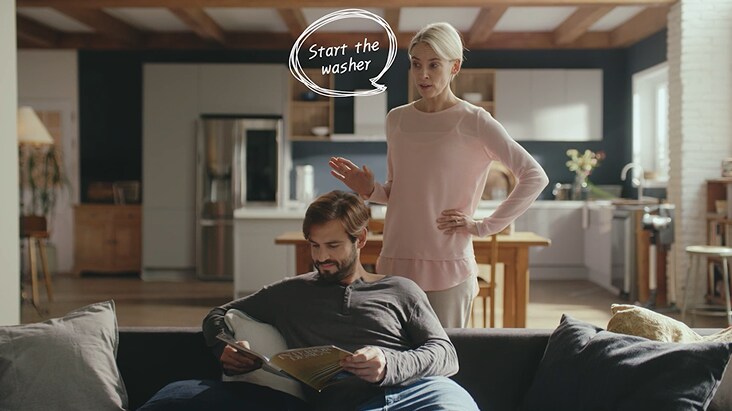 Dans une cuisine moderne et intelligente, une femme utilise une commande vocale pour démarrer la machine à laver avec LG ThinQ IA, tandis qu'un homme lit sur le canapé en arrière-plan.