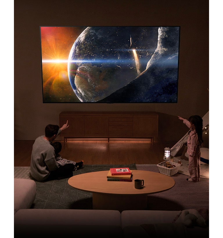 Une famille est assise sur le sol d’un salon faiblement éclairé, à côté d’une petite table, et regarde un LG TV fixé au mur, qui montre la Terre vue de l’espace.