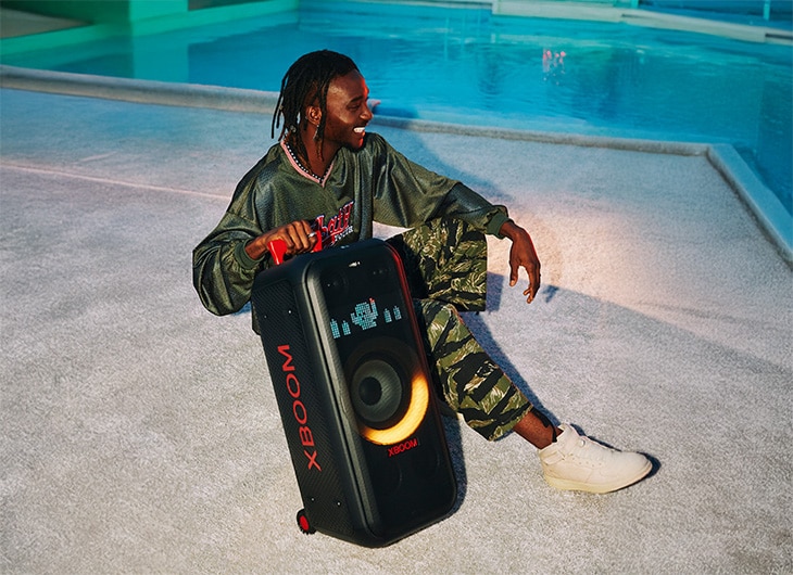 Persona relajándose junto a la piscina con la bocina portátil LG XBOOM, con iluminación naranja y un diseño elegante.
