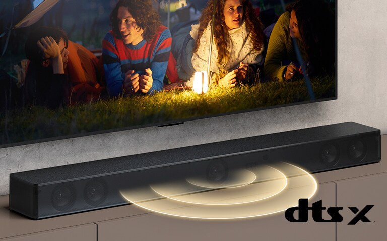 Un televisor LG en la pared, en la pantalla se muestran 2 parejas tiradas en el césped. Frente a ellos, hay una lámpara. La barra de sonido LG está debajo del televisor LG. Un gráfico de sonido sale del frente de la barra de sonido. El logotipo de DTS Virtual:X se muestra en la esquina inferior derecha de la imagen.