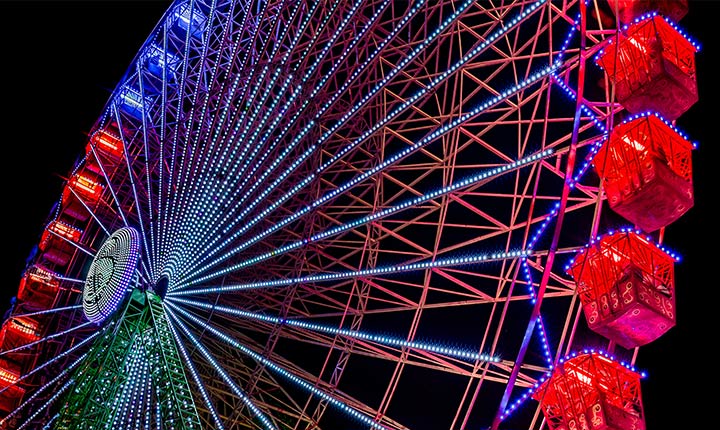 Una noria ligeramente descolorida se ilumina durante la noche, con destellos de colores que comienzan en el lado izquierdo de la pantalla y se mueven a través de ella. A medida que pasan los destellos de colores, la pantalla pasa de colores nublados a colores brillantes y vivos.
