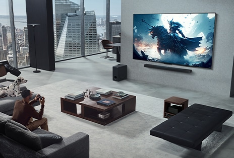 Una sala de estar con una persona jugando a un videojuego en su LG True Wireless TV y LG Soundbar montadas en la pared. Sostiene un controlador en la mano, pero la consola de juegos no se ve a simple vista. Tampoco se ve ningún cable. La Caja de conexión cero está oculta debajo de una mesa de apoyo y apenas puede verse.