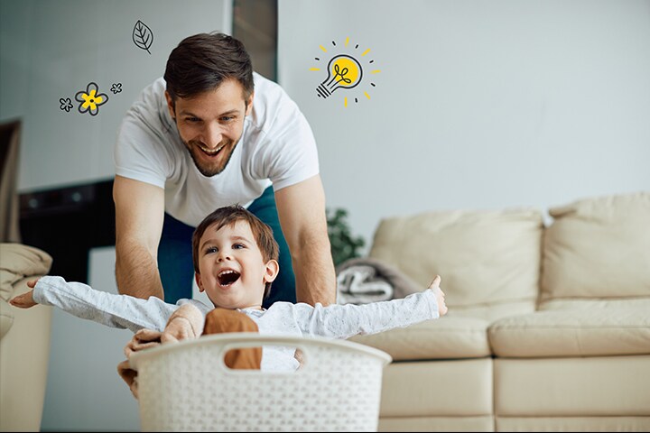 El padre empuja al niño hacia la cesta de la ropa sucia. A su alrededor hay bombillos que simbolizan el ahorro de energía, así como hojas y flores que simbolizan la naturaleza.