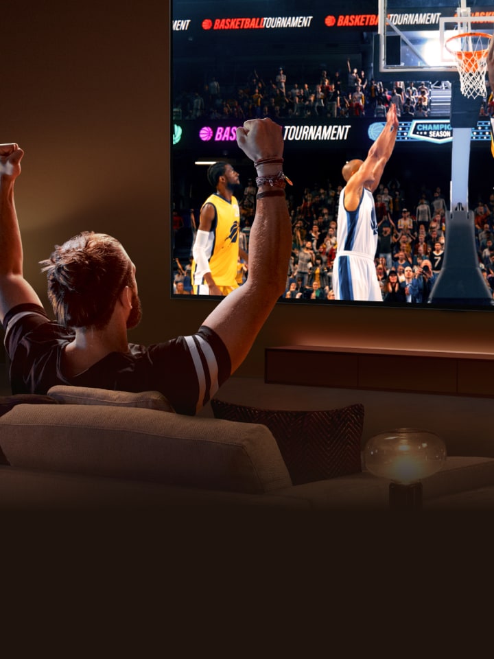 A man sits on a sofa in front of an ultra-large LG TV displaying a basketball game, with his arms in the air as if cheering.