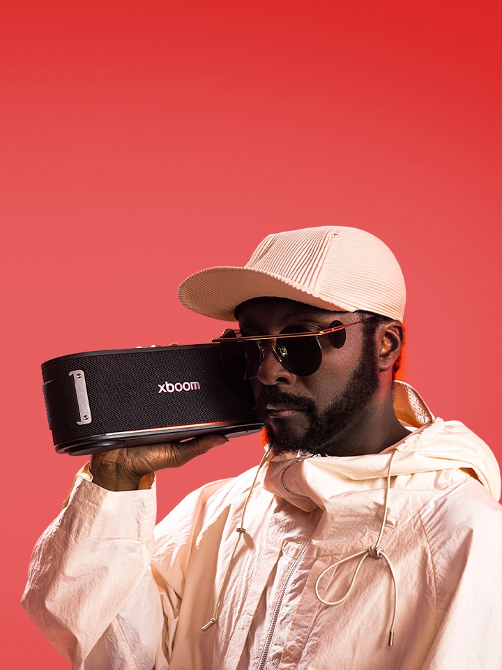 will.i.am in weißem Outfit und Sonnenbrille hält xboom Bounce direkt neben sein Gesicht.	