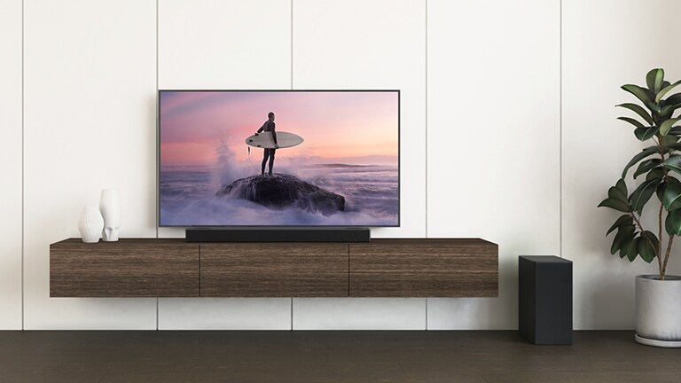 Ein LG TV und eine LG Soundbar stehen auf einem braunen Regal, und der Subwoofer steht auf dem Boden. Der Fernsehbildschirm zeigt einen Surfer, der auf dem Felsen steht.