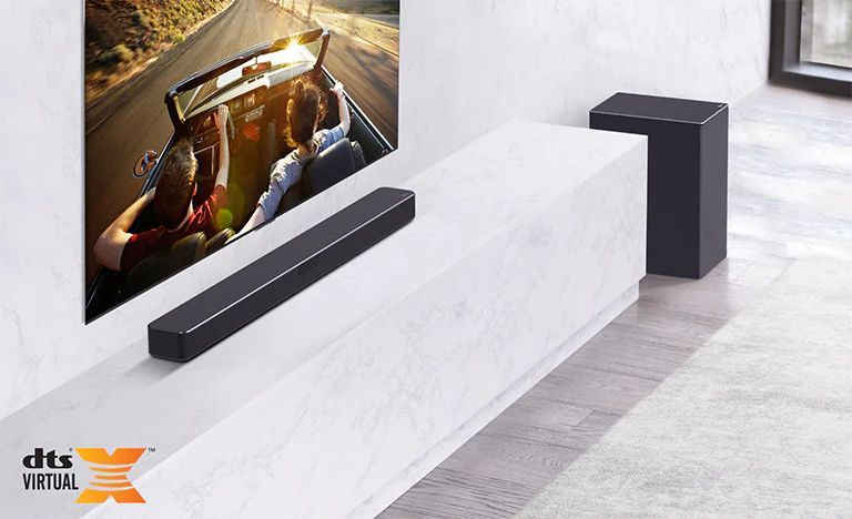 Der Fernseher hängt an der Wand, die LG Sound Bar steht unten auf einem Regal aus weißem Marmor mit einem Subwoofer auf der rechten Seite. Der Fernseher zeigt ein Paar in einem Auto.