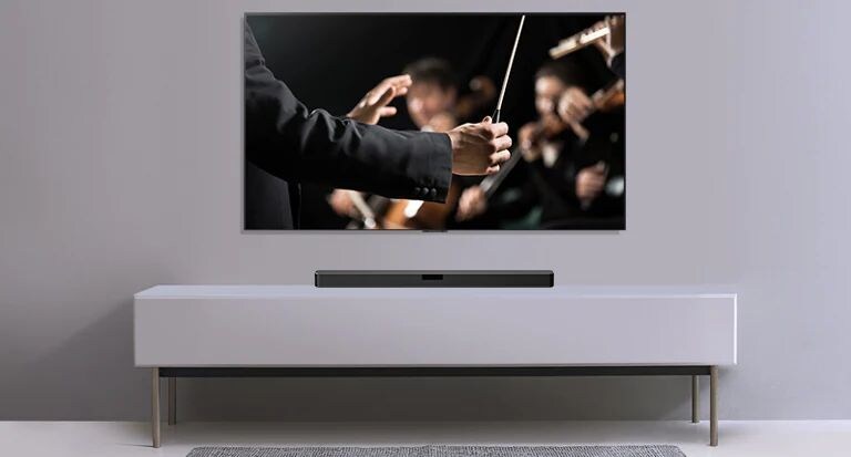 Vor einer grauen Wand wird ein Fernseher und darunter eine LG Sound Bar auf einem grauen Regal gezeigt. Der Fernseher zeigt einen Dirigenten, der ein Orchester dirigiert.