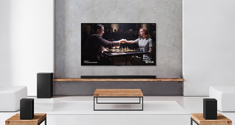 Eine Zusammenstellung, bestehend aus zwei Rücklautsprechern, Subwoofer, einer Soundbar sowie einem TV, befindet sich in einem hellen Wohnzimmer. Ein Poster einer TV-Sendung ist auf dem Bildschirm zu sehen.