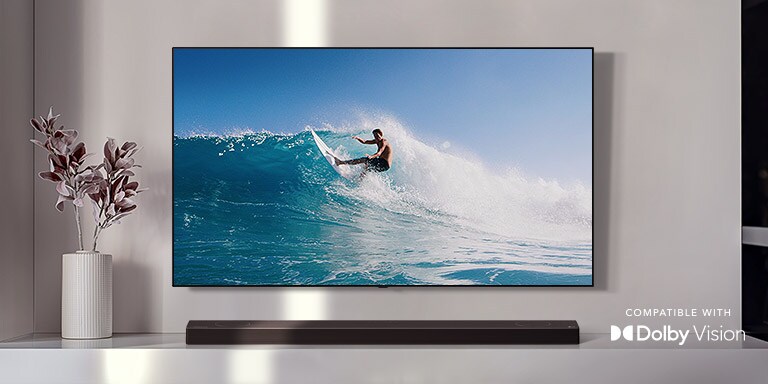 An der Wand befestigter TV. Der TV zeigt einen Mann, der auf einer großen Welle surft. Die LG Soundbar steht direkt unter dem TV auf einem weißen Regal. Direkt neben der Soundbar befindet sich eine Vase mit einer Blume (Video abspielen).