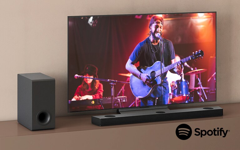 Ein LG TV zeigt ein Konzert und die LG Soundbar befindet sich unter dem Fernseher. Links steht der Subwoofer auf einem braunen Regal.
